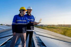 Two-workers-in-blue-uniforms-with-yellow-and-white-hard-hats-using-laptop-near-solar-panels