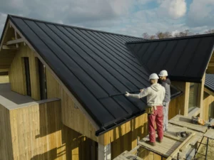 two-construction-workers in-safety-gear are-installing a-black-metal-roof on-a-wooden-house.