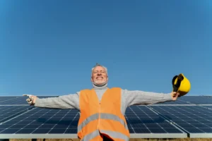 Worker-in-orange-vest-holding-yellow-hard-hat-in-front-of-solar-panels