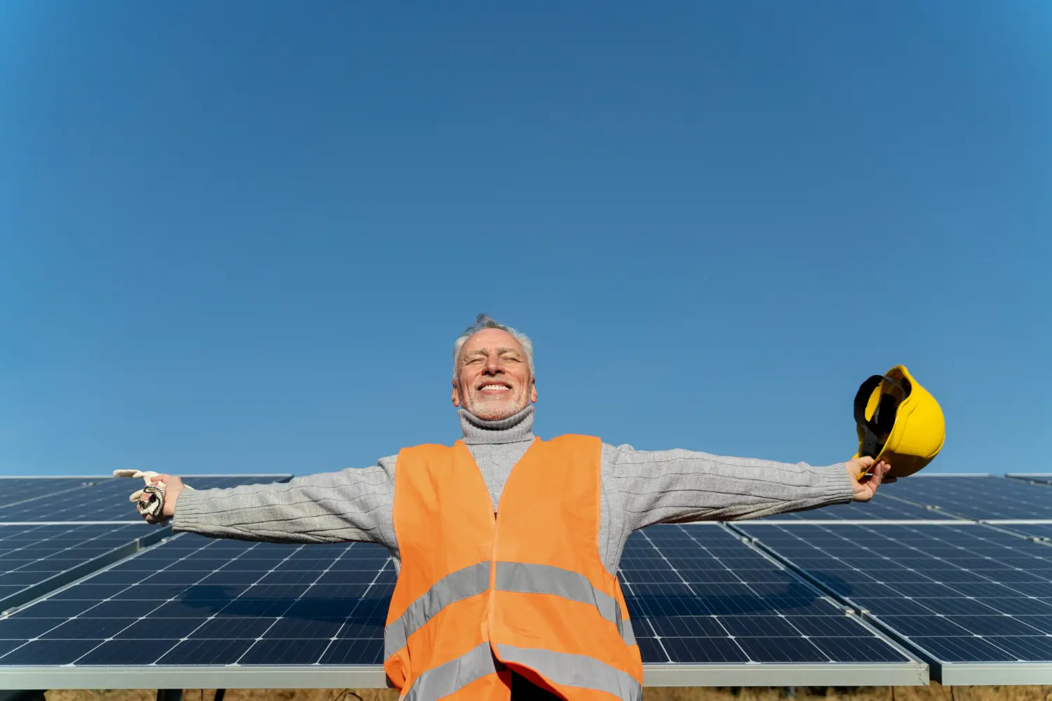 Worker-in-orange-vest-holding-yellow-hard-hat-in-front-of-solar-panels